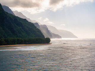Lush green cliffs of the Napali Coast descend into the Pacific Ocean, with mist, sunlight, a sandy...