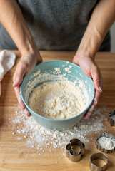 A person is holding a blue bowl filled with flour, with cookie cutters and flour scattered on a wooden surface nearby