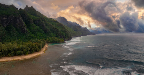 Aerial view of Kauai's Napali Coast with jagged green cliffs, a sandy beach, turquoise waters, lush vegetation, and a vibrant sky with dramatic clouds.