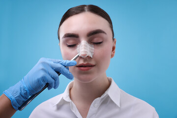 Doctor removing medical bandage from patient's nose after plastic surgery operation on light blue background, closeup