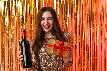 Happy young woman with gift box and bottle of wine against foil curtain