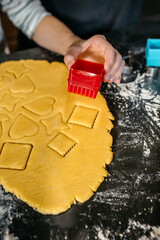 Unrecognizable young child hand using cookie cutters of various shapes on rolled out dough on kitchen counter, with a rolling pin nearby, preparing for baking. Siblings making festive cookies together