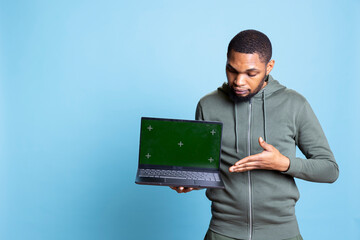 Jolly african american person presenting green screen display on laptop, doing recommendation. Friendly young guy pointing at mockup on notebook, wearing casual clothes in studio.