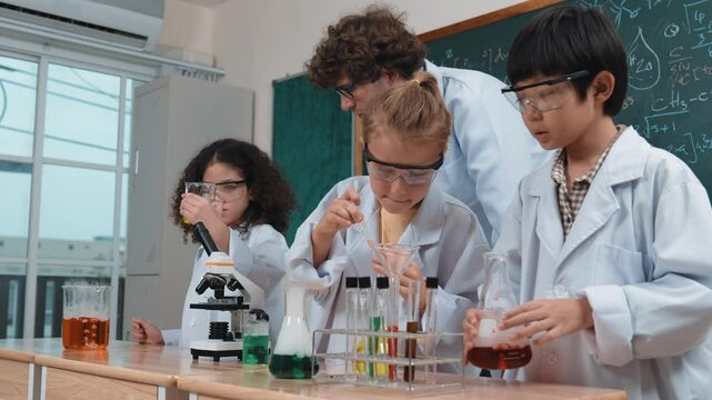 Caucasian boy drop chemical solution in example while diverse student doing experiment. American girl inspect colored liquid while standing with teacher looking after children at blackboard. Pedagogy.