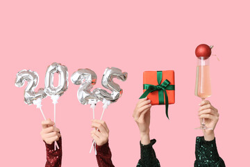 Female hands with figure 2025 made of foil balloons, gift box, Christmas ball and glass of champagne on pink background