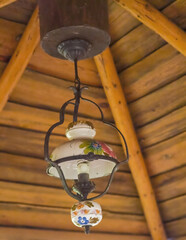 Stara, zabytkowa porcelanowa lampa elektryczna (przerobiona z lampy naftowej) zwisa z sufitu w drewnianej altanie. Zabytkowa porcelanowa lampa elektryczna, pierwotnie lampa naftowa,  ma kwiatowe wzory © Grzegorz