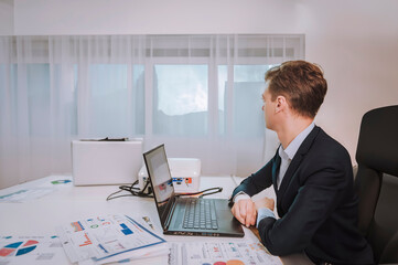 A young business man in an office, working on a laptop at a desk, thinking about something, and looking out the window