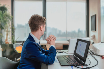 A young business man in an office, working on a laptop at a desk, thinking about something, and looking out the window