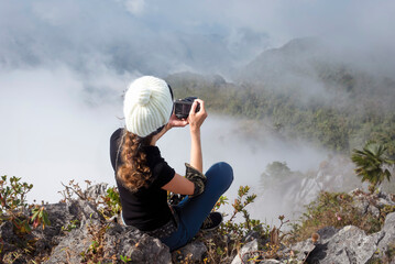 A young girl sitting on a rock while hiking in the mountains and snapping pictures