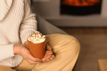 Beautiful young woman with cup of hot chocolate and whipped cream sitting in evening at home, closeup