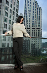 Confident woman in a cozy knit sweater and black pants poses on a balcony with a modern cityscape. A chic and urban winter fashion portrait for lifestyle, editorial, and advertising use.