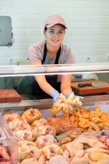 Young woman butcher in apron lays out raw chicken meat on counter in butcher shop