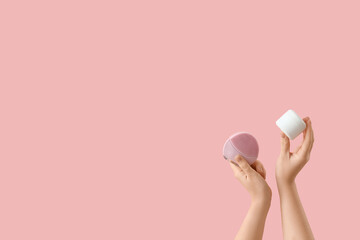 Female hands with jar of cream and silicone facial cleansing brush on pink background