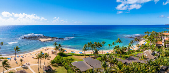Aerial view of a crescent shaped beach on Kauai, Hawaii, with turquoise waters, palm trees, resort buildings, and visitors under umbrellas.