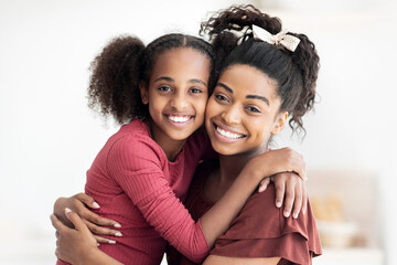 Closeup portrait of happy black mother and daughter hugging and smiling at camera, beautiful young african american woman mom and teen girl kid cuddling at kitchen, copy space