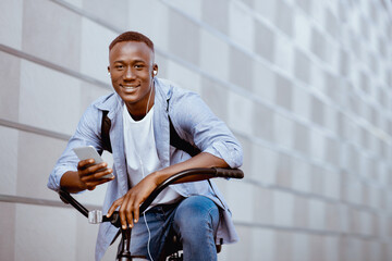 Handsome black guy with rucksack, earphones and mobile device riding bicycle near brick wall...