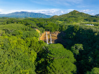 Twin waterfalls cascade into a green basin surrounded by dense tropical vegetation, with rolling hills and mountains under clear skies in Kauai, Hawaii.