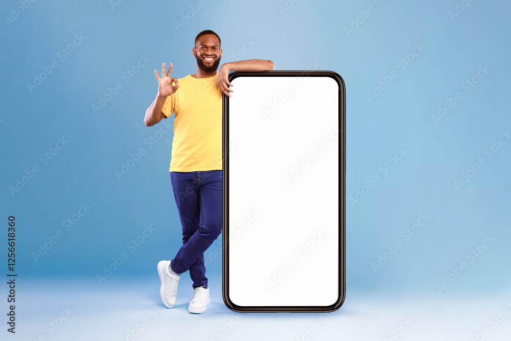 Poster positive african american guy showing okay gesture next to huge smartphone on blue studio background