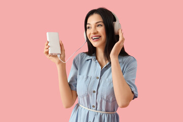 Beautiful young woman with modern power bank and headphones on pink background