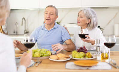 Two elderly women and elderly man talking and drinking wine in kitchen