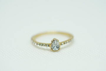 Close-up of a delicate gold ring with a teardrop-shaped clear gemstone and side stones, placed on a white textured background. Perfect for bridal, fashion, elegance, and luxury jewelry themes.