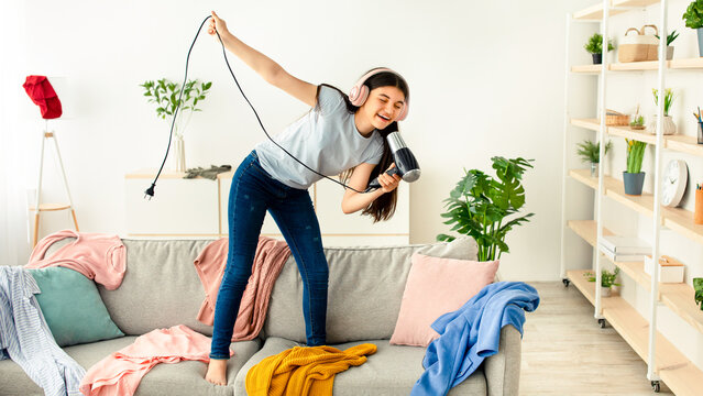 Cheerful Indian teen girl with blowdryer as mic singing her favorite song and dancing on sofa at home, full length. Cool teenager pretending to be famous singer, having fun in messy room