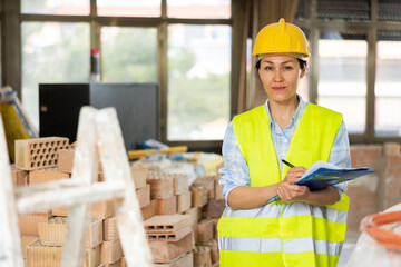 Woman foreman in a protective helmet and yellow vest checks the completed construction work on the drawing