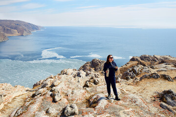 Fototapeta premium A woman traveler stands on a rock with a thermos with hot tea in her hands and enjoys the beautiful landscape. Lake Baikal in spring. Ice melting time. The concept of freedom and travel.