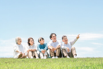青空の見える公園・丘に座る家族・親子・三世代家族（父親・母親・娘・祖母・祖父）
