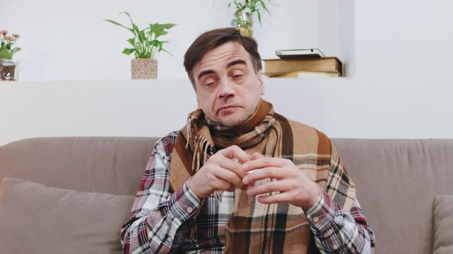 An elderly man, wrapped in a cozy blanket, sits on a sofa surrounded by tissues, sneezing, coughing and fighting off a cold in a bright living room filled with plants and cozy decor.