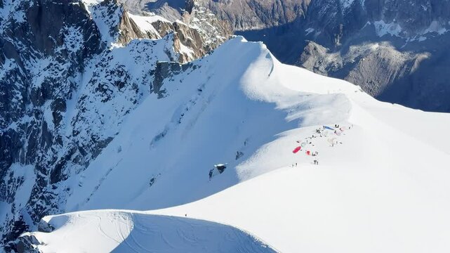Paragliders with backpacks descend the steep snowy slopes of Aiguille du Midi, Chamonix, preparing for a parachute jump between the majestic peaks of the French Alps, with amazing view of Mont Blanc