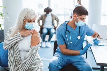 I Got My Coronavirus Vaccine. Portrait Of Older Woman Patient In Medical Face Mask Showing Vaccinated Arm After Antiviral Injection For flu Protection, Looking At Her Shoulder With Sticking Patch