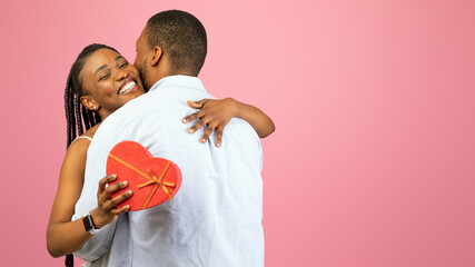 Excited black woman holding heart shaped gift box and hugging her boyfriend on pink studio background. Lovely African American couple celebrating Women's or Valentine's Day together, free space