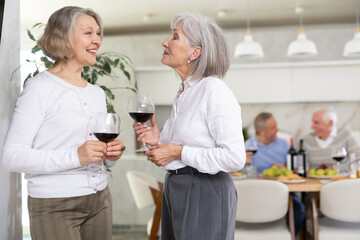 Two elderly women friends chatting and drinking wine in kitchen at home