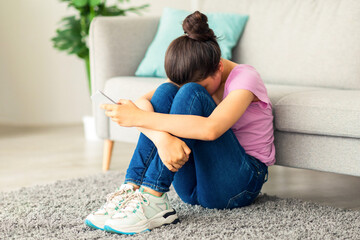 Depressed Indian teen girl with cellphone crying on floor at home, suffering from cyber bullying,...