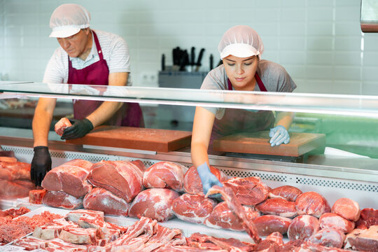 Adult man and young woman butchers in aprons put raw meat beef in butcher shop window - Powered by Adobe