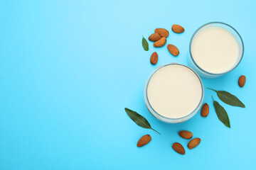 Fresh nut milk in glasses, almonds and green leaves on light blue background, flat lay. Space for text