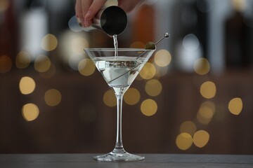 Woman making tasty martini cocktail with olives at wooden table on blurred background, closeup. Bokeh effect