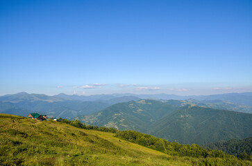 Obraz premium Tranquil mountain landscape showcasing a house, by a green forest with rolling hills and peaks in the background bathed in sunlight under a beautiful blue sky. Carpathians, Ukraine