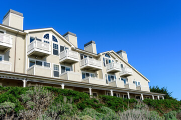 Modern seaside residential apartments building with beige siding, white balconies, and arched windows, surrounded by lush greenery under a vibrant blue sky