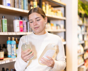 Young woman housewife in household store examines package, chooses shampoo. Customer reads label, compare varieties items. Buyer is shopping in household department.