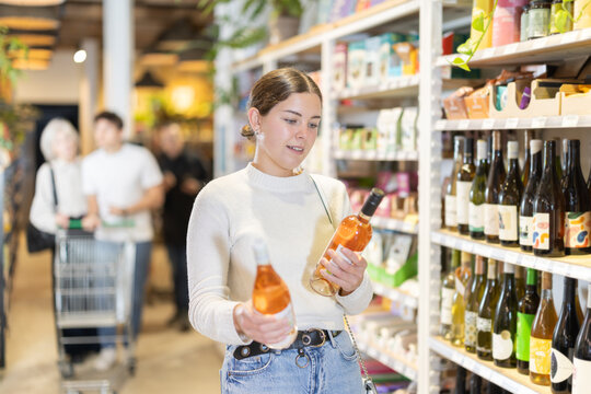 Young woman holds bottles of wine in her hands, reading labels and stands near the shelves with alcohol in a hypermarket against the background of customers.