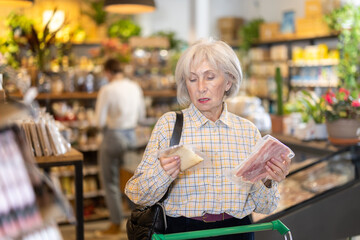 Mature woman with gray hair in an eco supermarket chooses a package of cheese and bacon. Senior...