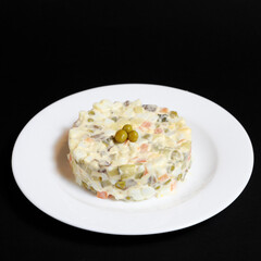 Serving of Olivier salad on a white plate, shot on a black background, topped with three green pickled peas