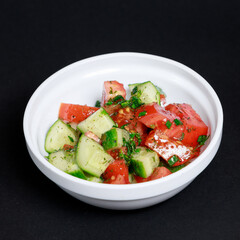Fresh cucumber and tomato salad with chopped dill and green onions in a white bowl, shot on a black background
