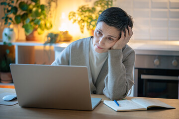 Fototapeta premium Fatigued distracted woman leans on hand while working on laptop late at home, struggle to maintain focus. Overwork burnout, lack of time, sleep deprivation, headache, depression, emotional strain