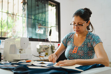 Mid-age latin hispanic woman working in a sewing workshop. Using white chalk to trace sewing pattern in dark fabric. Fashion designer concept.