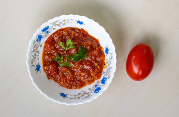 Delicious homemade Tomato Bhorta served in a white bowl atop a light wooden surface. A whole red tomato rests nearby. Tomato bhorta is a popular Bengali dish. Top view