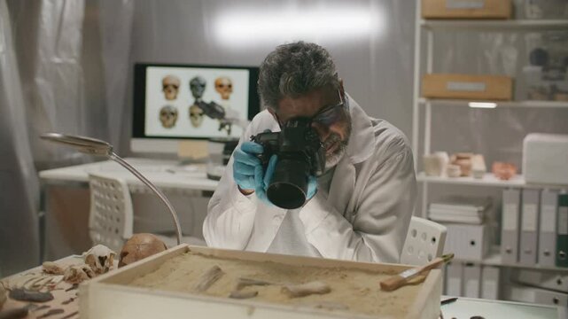 Senior archaeologist in lab coat and gloves photographing ancient bones and artifacts in research laboratory, using professional camera for documentation and analysis