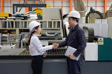 Two worker shake hands in a factory. They wear safety helmet stand in front of High Precision Fiber...